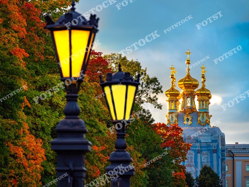 На переднем плане фотографии видны старинные фонари, освещающие осеннюю аллею в Екатерининском парке Царского Села. Вдалеке возвышаются золотые купола дворцовой церкви Воскресения Христова, отражающие последние лучи вечернего света. Это сочетание архитектуры, осенней природы и мягкого света фонарей придает композиции атмосферу уюта и спокойствия. Екатерининский дворец и его парк являются одним из наиболее известных и посещаемых мест Санкт-Петербурга.